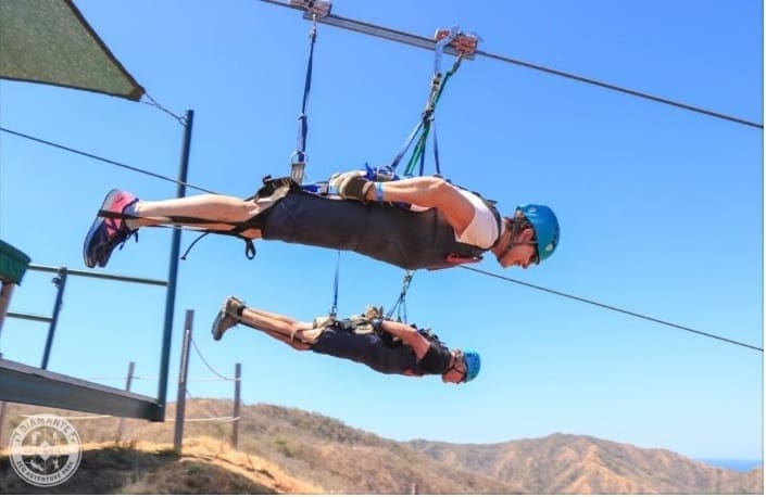 Two people wearing helmets and safety harnesses are ziplining on a high ropes course outdoors, with hills in the background.