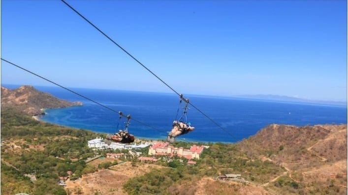Two people ride a zip line over a rugged coastal landscape with the blue ocean in the background.