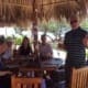 Crusaders Rugby Club members sit around a wooden table under a thatched canopy outdoors, smiling and giving thumbs up.