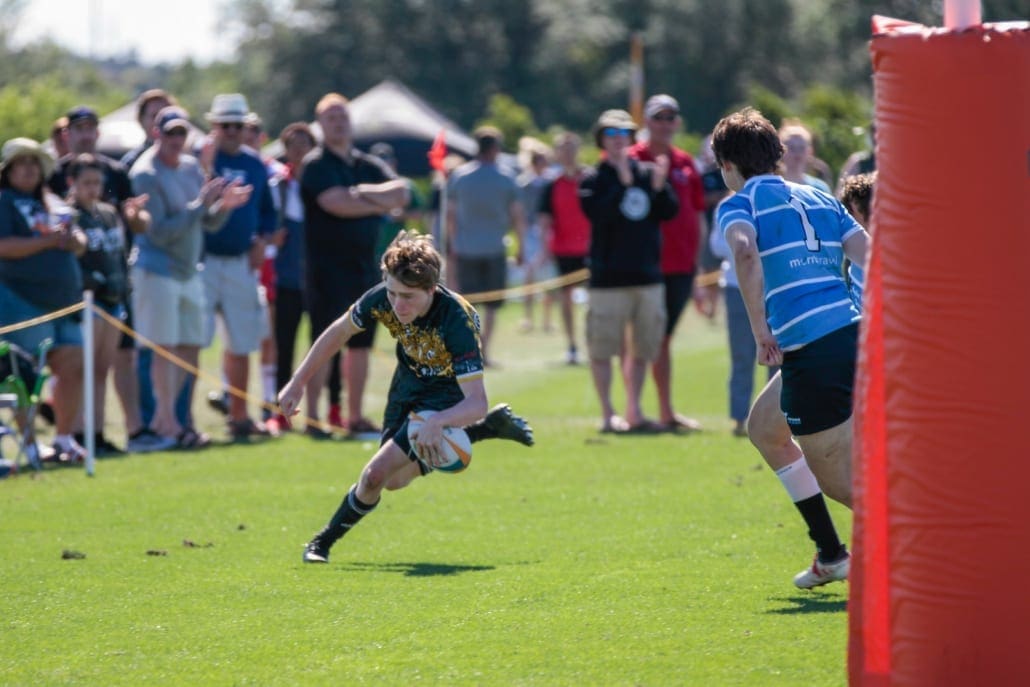 Young rugby player in black and yellow sprints with the ball past a blue-jersey defender on a sunny field, with spectators and a red padded goal post in the background.