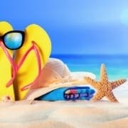 Yellow flip-flops with sunglasses sit on the sandy beach beside a starfish, with the blue ocean in the background.