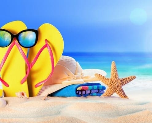Yellow flip-flops with sunglasses sit on the sandy beach beside a starfish, with the blue ocean in the background.