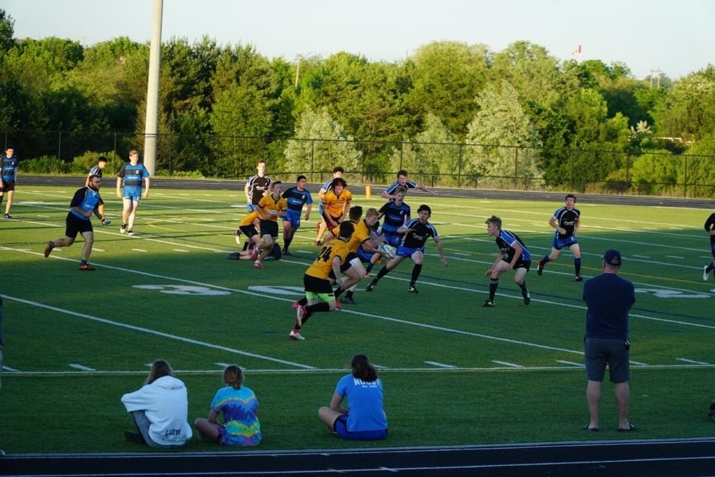 ORG_DSC09333 Crusaders Rugby Club players in yellow and blue uniforms compete on a grassy rugby field in Oakville with spectators along the sideline.