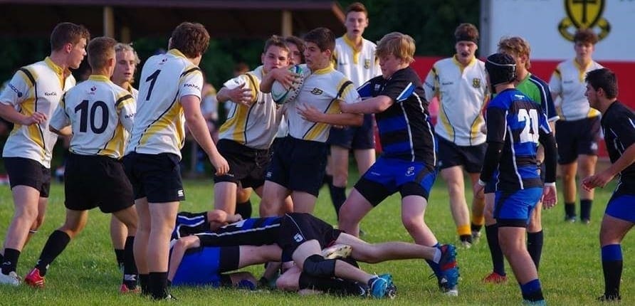 Aurora1 Crusaders Rugby Club players in white and gold jerseys gather around a tackled player on a grassy field during a match in Oakville.