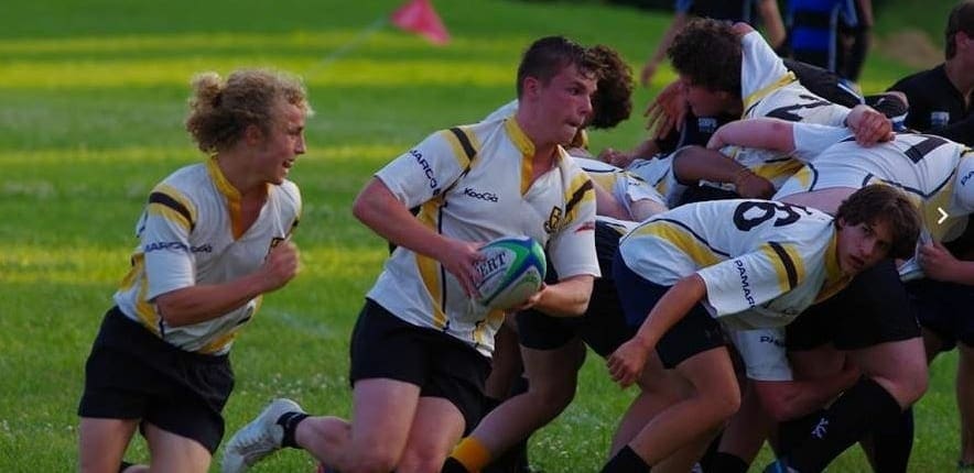 Aurora5 Crusaders Rugby Club players in white jerseys with gold trim chase the ball during a match on a grassy field.