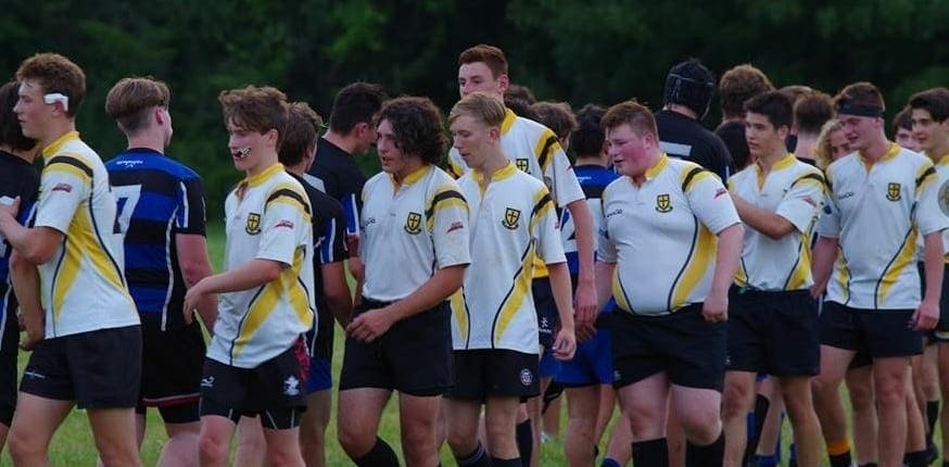 Aurora7 Crusaders Rugby Club players stand in a line on a grassy field wearing white and yellow jerseys.