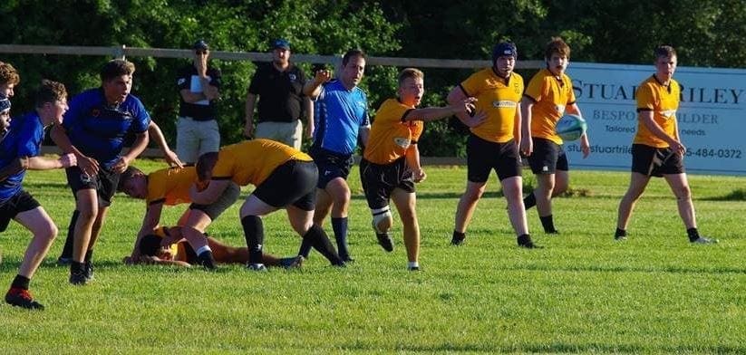 MT5 Crusaders Rugby Club players in blue and yellow jerseys engage in a rugby scrum on a sunny grassy field in Oakville.