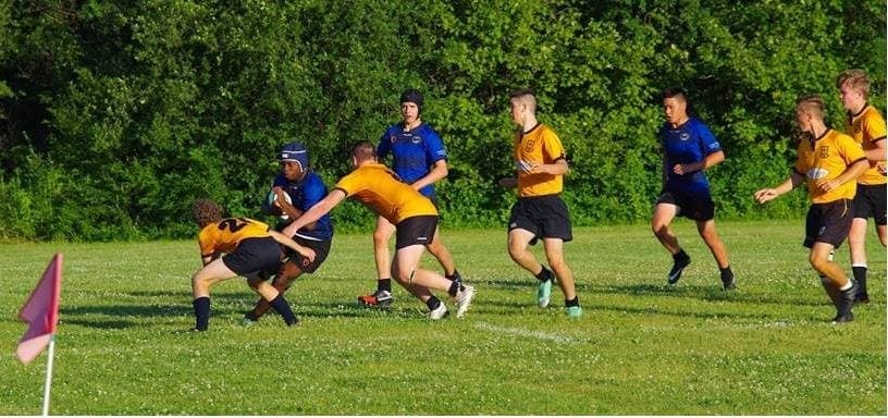 MT6 Rugby players in yellow and blue jerseys contest the ball on a sunny grassy field near a corner flag.