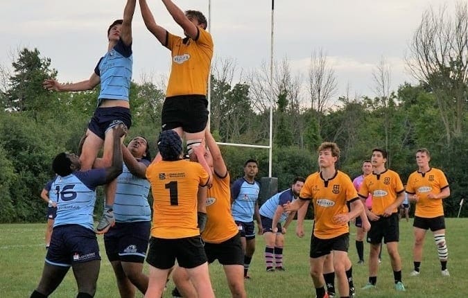 Rugby players in orange and light-blue jerseys contest a lineout on a grassy field.