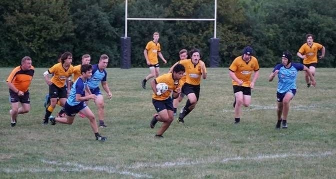 Crusaders Rugby Club players in orange and blue jerseys play a match on a grassy field, with a ball carrier in the foreground.