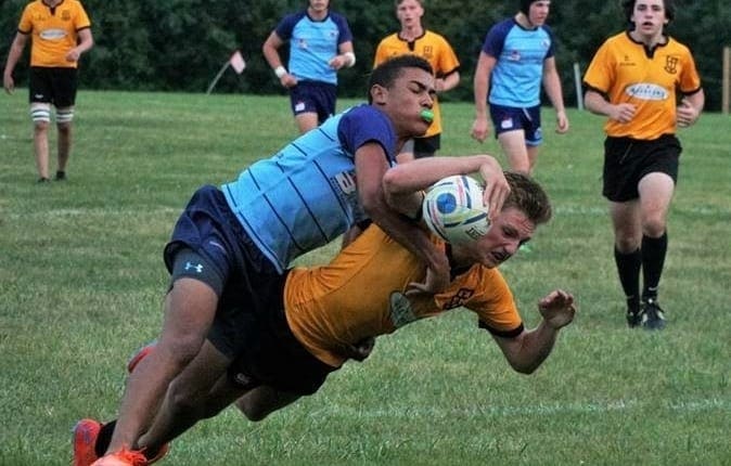 Crusaders Rugby Club players tackle on a green field as a yellow-shirted player carries the ball.