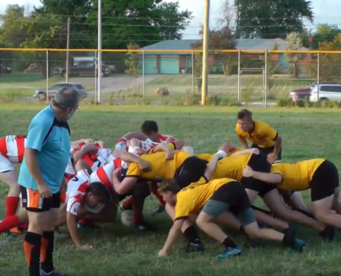 Rugby players in a scrum on a grassy field wearing red-and-white striped and yellow jerseys at Crusaders Rugby Club in Oakville.
