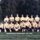 Vintage team photo of a rugby club in yellow jerseys posing in two rows on a grassy field.