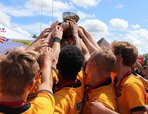 Crusaders Rugby Club players in yellow jerseys huddle on the field, raising a trophy under a blue sky.