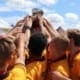 Crusaders Rugby Club players in yellow jerseys huddle on the field, raising a trophy under a blue sky.