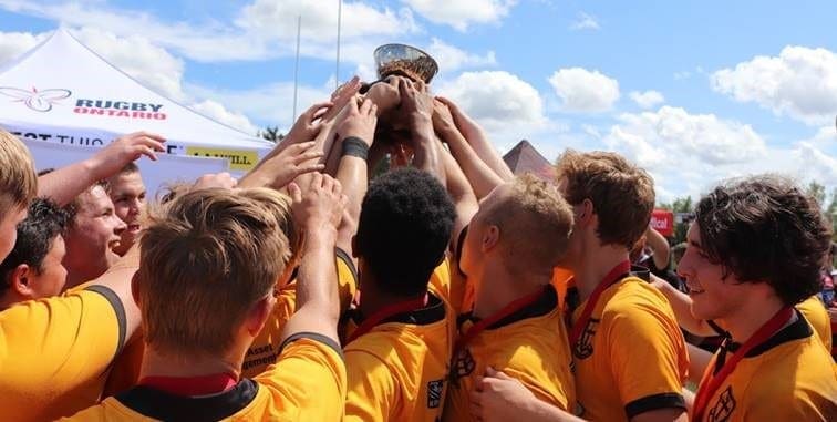 Crusaders Rugby Club players in yellow jerseys huddle on the field, raising a trophy under a blue sky.