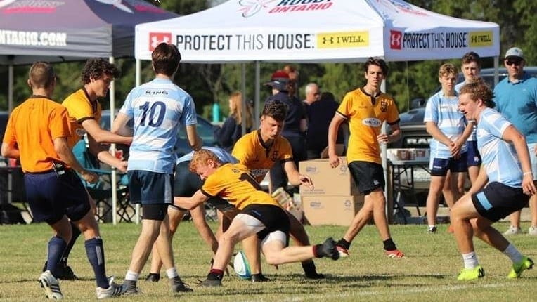 Crusaders Rugby Club players in orange and blue compete on a grassy field with tents and banners in the background.