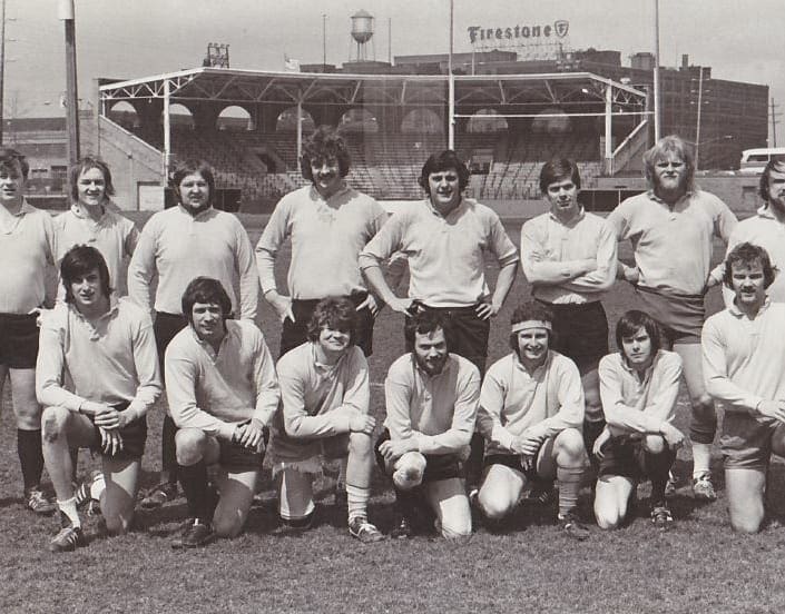 A black and white team photograph captures thirteen men in athletic attire posing on a grassy field with a stadium displaying a Firestone sign in the background.