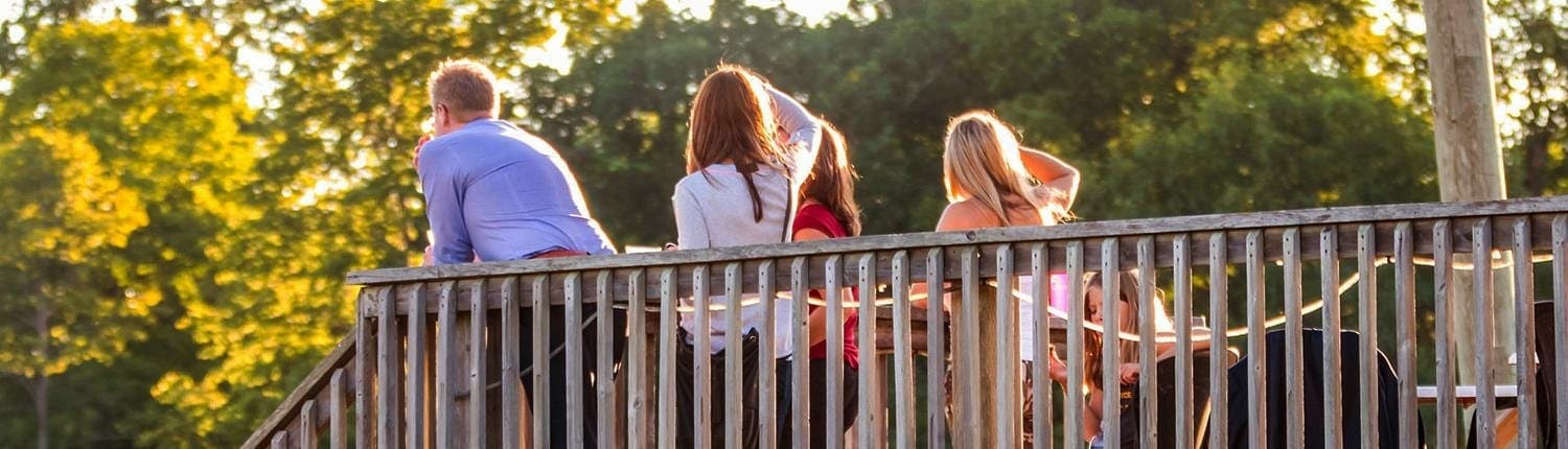 A group of people leaning on a wooden railing outdoors with trees and sunlight in the background.