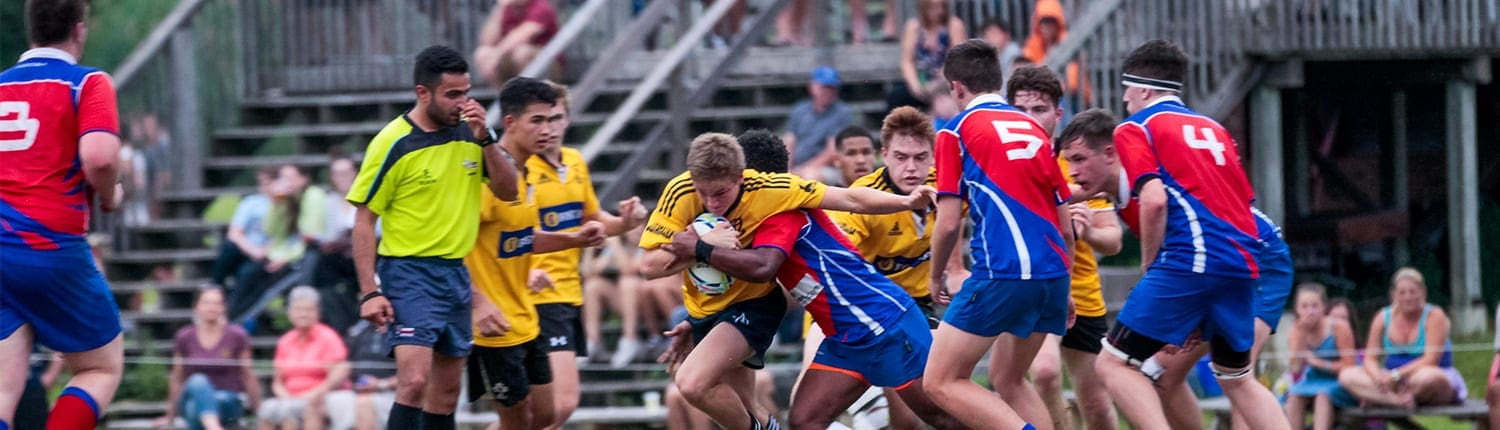 Crusaders Rugby Club Oakville players in red jerseys and blue shorts clash on the field near the sideline, with a yellow-clad referee and spectators in the stands. Crusaders Rugby Club Oakville players in red jerseys and blue shorts clash on the field near the sideline, with a yellow-clad referee and spectators in the stands.