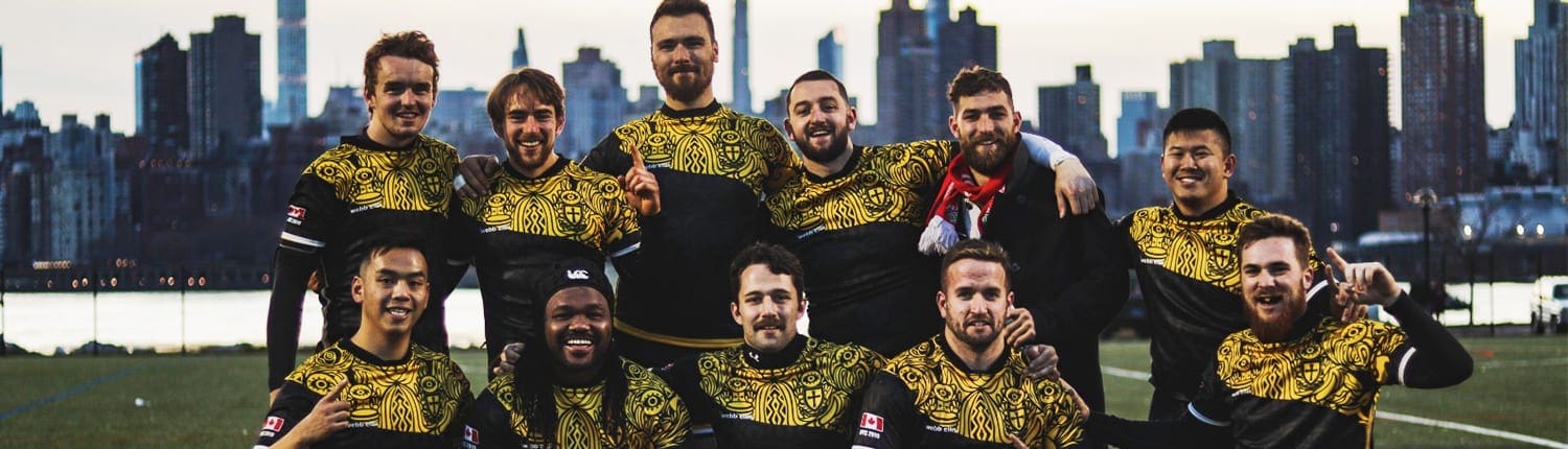 Group of Crusaders Rugby Club players in yellow and black uniforms pose for a team photo on a rugby field with the Oakville skyline in the background.