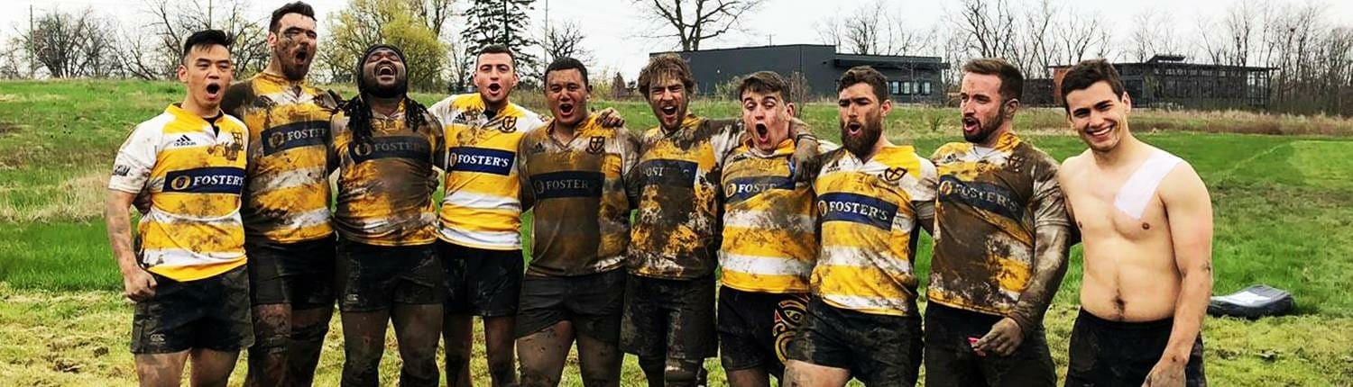 Group of muddy Crusaders Rugby Club players posing on a grassy field in yellow and black striped jerseys in Oakville. Group of muddy Crusaders Rugby Club players posing on a grassy field in yellow and black striped jerseys in Oakville.