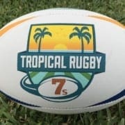 Rugby ball on grass featuring the Tropical Rugby logo with palm trees and a 7s badge.