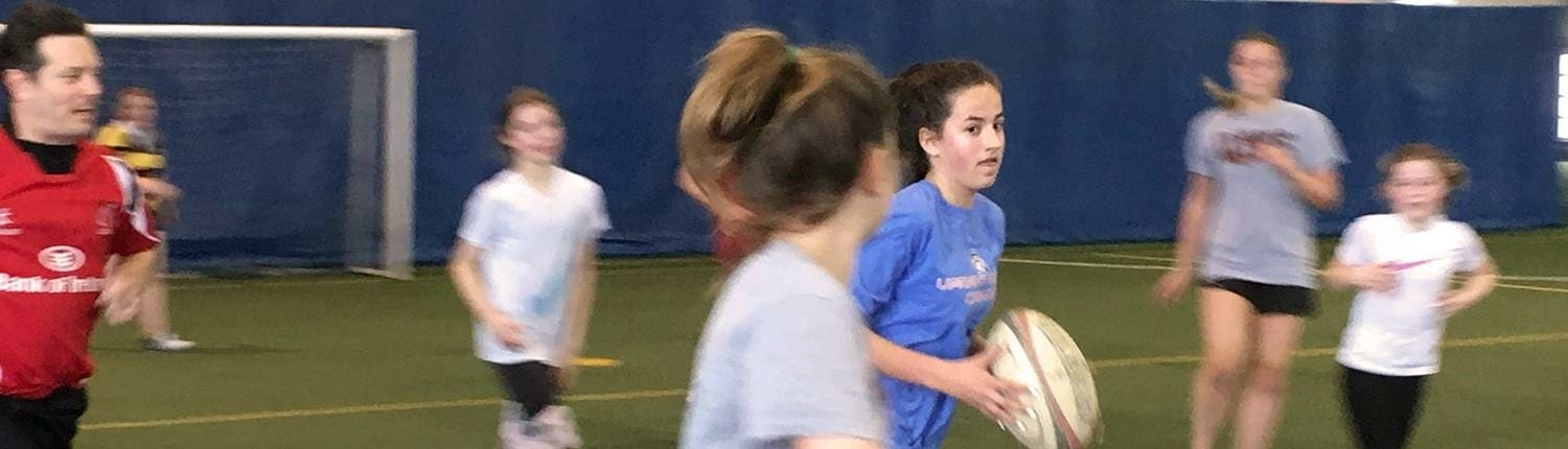 Young Crusaders Rugby Club players practice on an indoor field, with a girl in a blue jersey holding a rugby ball. Young Crusaders Rugby Club players practice on an indoor field, with a girl in a blue jersey holding a rugby ball.