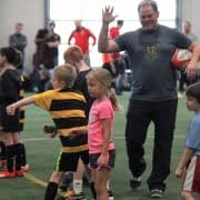 Crusaders Rugby Club Oakville coach raises his hand while guiding young players on an indoor turf rugby field.