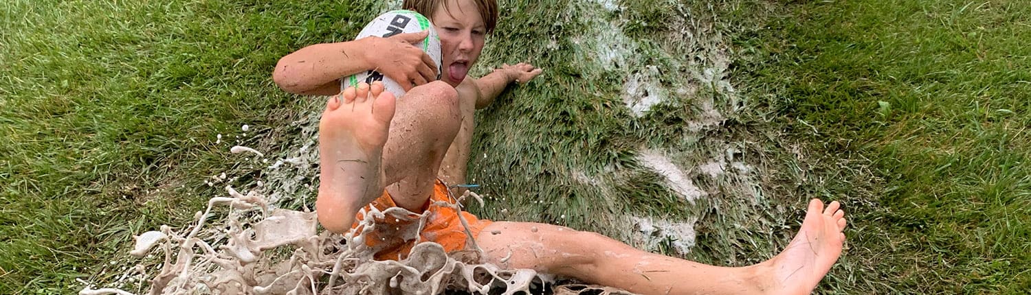 A young boy in orange shorts sits barefoot in mud beside a tree outdoors. A young boy in orange shorts sits barefoot in mud beside a tree outdoors.
