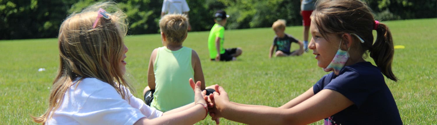 Two young girls hold hands during a rugby training session on a sunny grassy field with other players in the background. Two young girls hold hands during a rugby training session on a sunny grassy field with other players in the background.