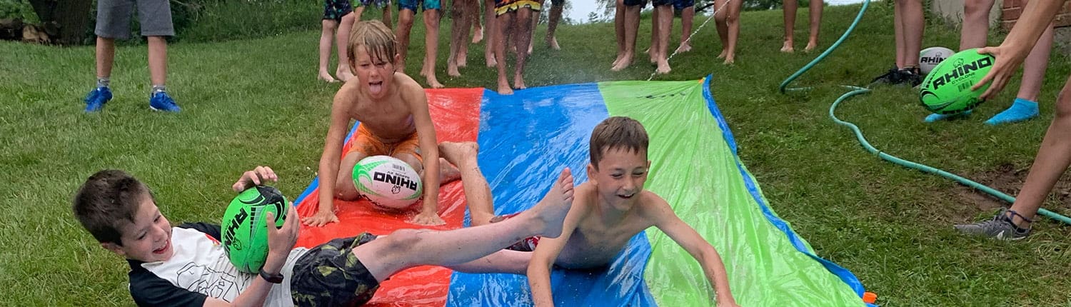 Young Crusaders rugby players slide down a blue and green inflatable slide on a grassy field, with a rugby ball visible. Young Crusaders rugby players slide down a blue and green inflatable slide on a grassy field, with a rugby ball visible.