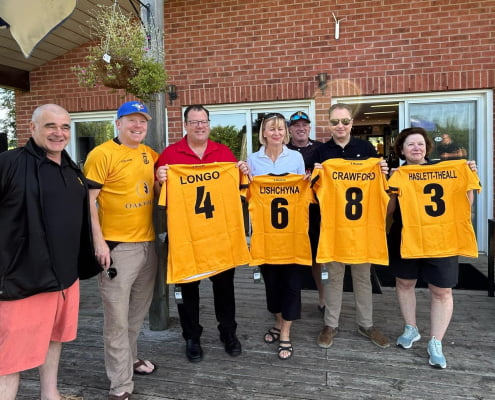 Seven adults on a patio hold yellow sports jerseys with names and numbers, smiling.