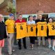 Seven adults on a patio hold yellow sports jerseys with names and numbers, smiling.