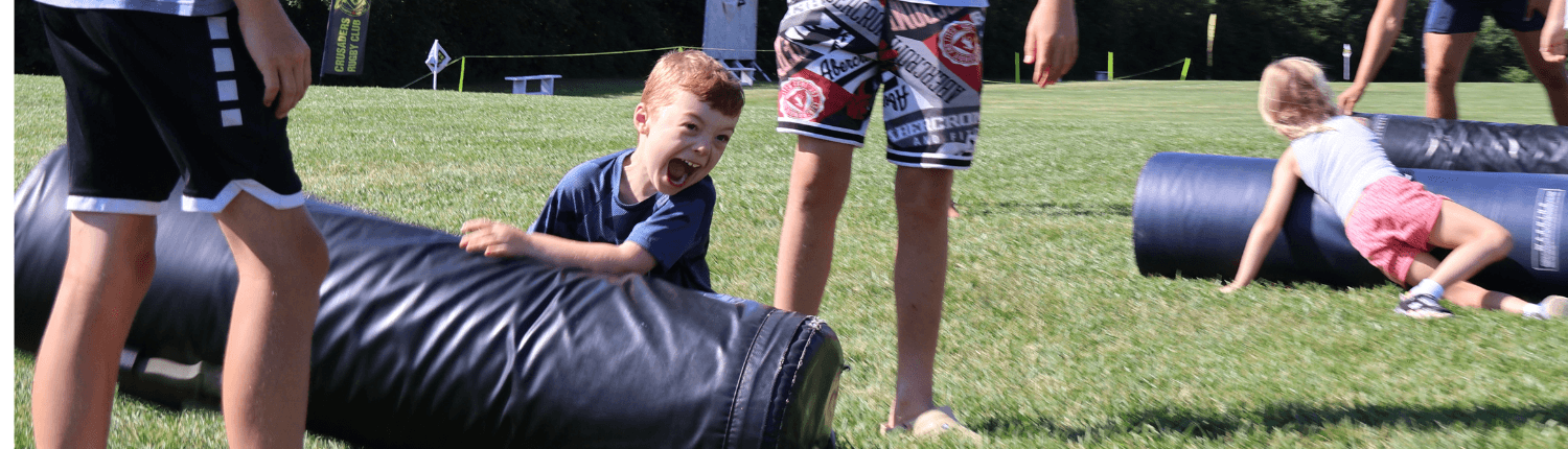 Children and coaches on a grassy rugby field at Crusaders Rugby Club in Oakville, with large black training cylinders in the foreground.