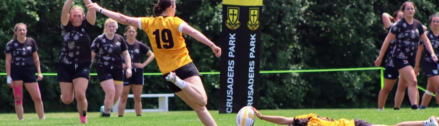 Crusaders Rugby Club players in Oakville play a rugby match on a grassy field, with a yellow-jersey player racing past black-jersey opponents.