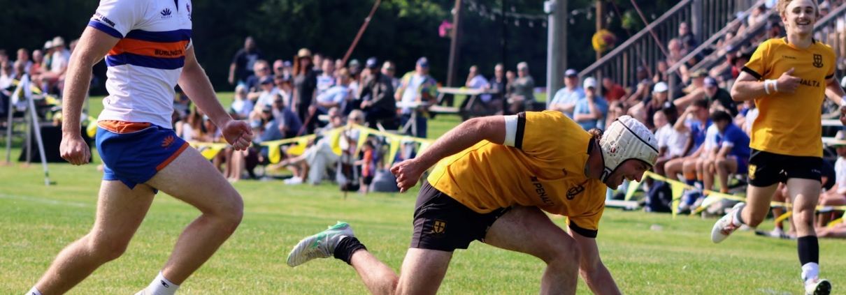 Hurling player in yellow jersey and white helmet scoops ball on sunny field, spectators watching.