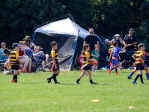Kids flag rugby game on sunny green field with colourful jerseys, coaches, tent, and trees.