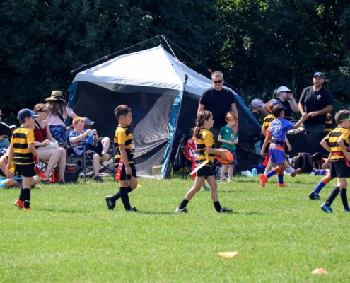 Kids flag rugby game on sunny green field with colourful jerseys, coaches, tent, and trees.