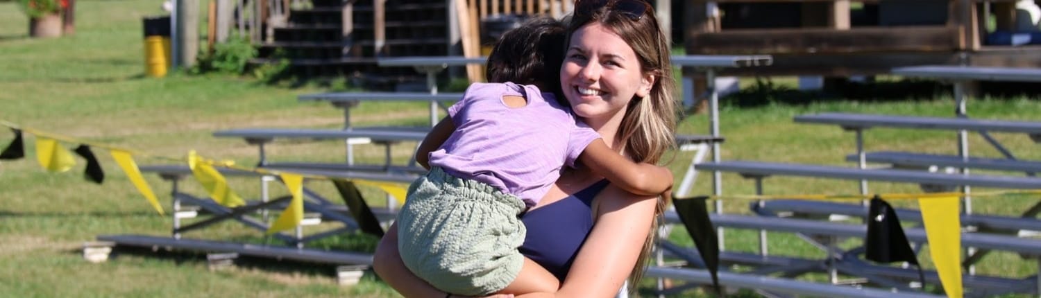 A smiling woman hugs a child on a grassy rugby field at Crusaders Rugby Club in Oakville, with training equipment in the background.