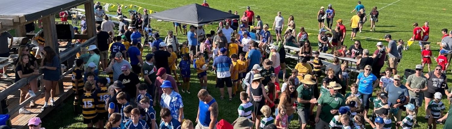 Minis Tournament A large crowd of players, coaches and spectators gathers on a sunny grassy rugby field at Crusaders Rugby Club in Oakville, with tents and viewing areas in the background.