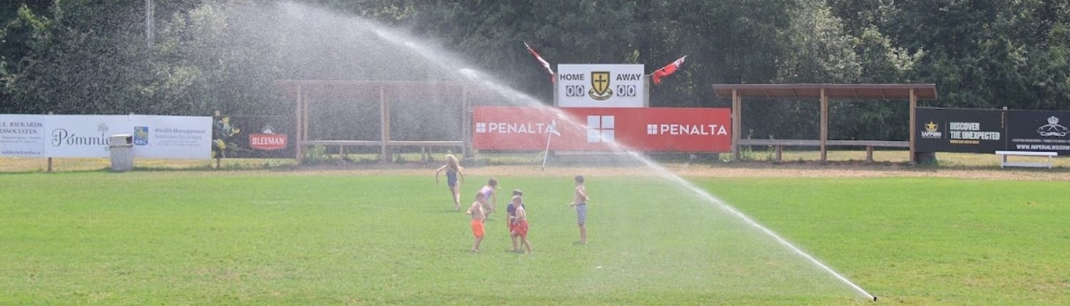 Summer Camps Crusaders Rugby Club players on a green rugby field in Oakville, with sprinklers watering the pitch and banners in the background.