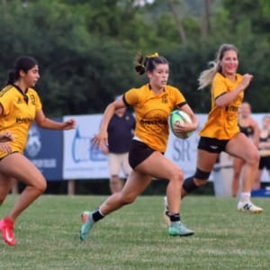 U18G-2025 Three female rugby players in yellow jerseys sprint across a grassy field during a match, with one player carrying the ball.