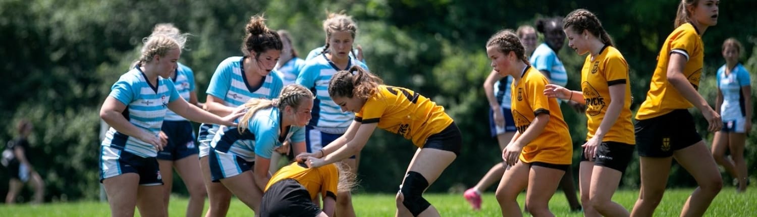 U18 Girls Rugby players from the Crusaders Rugby Club in Oakville compete on a grassy field, wearing blue-and-white striped and yellow jerseys.