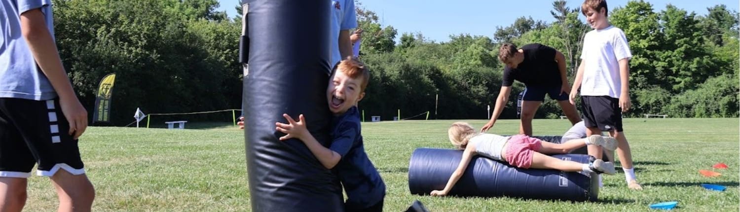 Young rugby players train on a sunny grass field with a blue tackle shield and training cones.